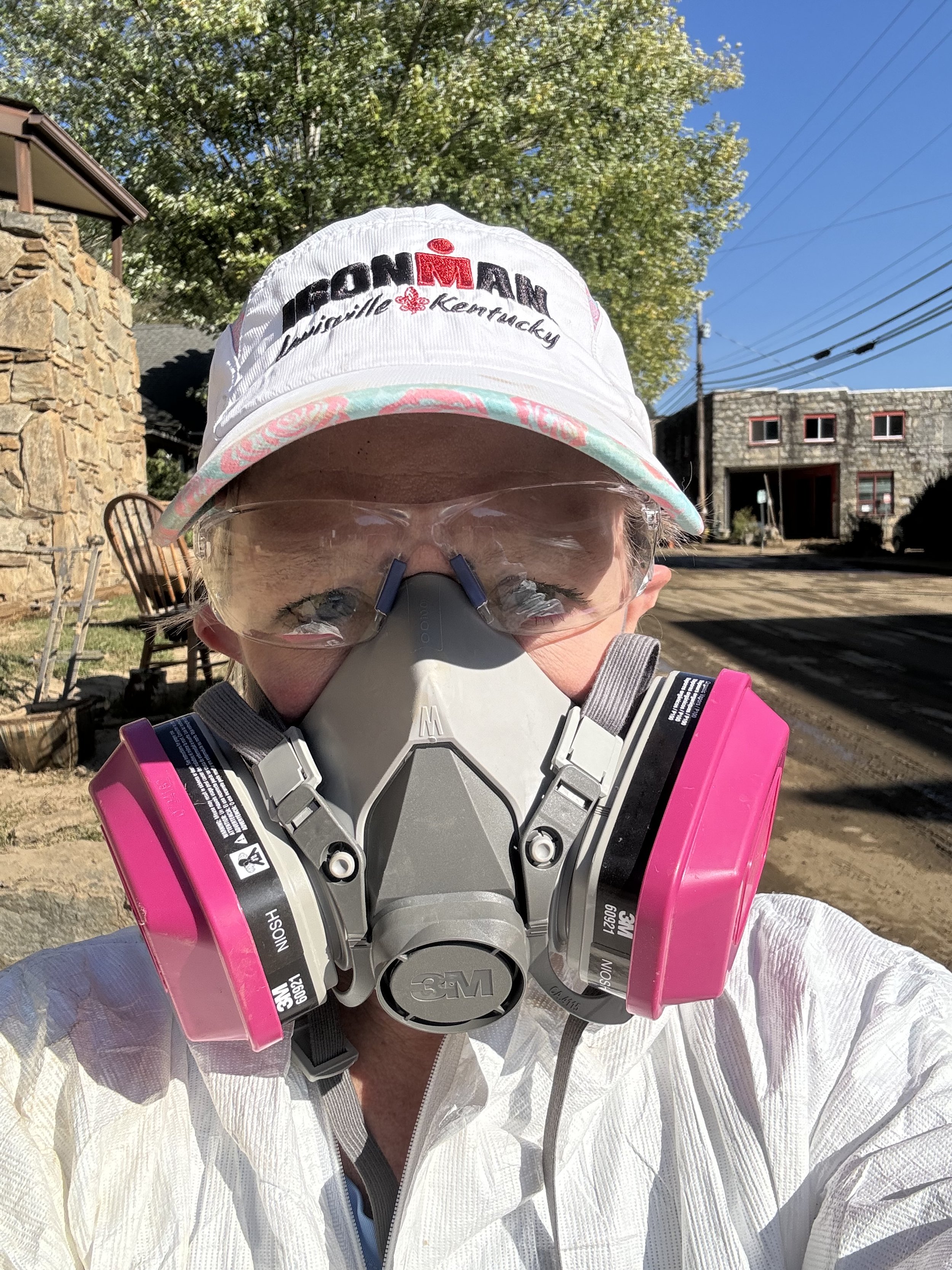Person wearing a white sun hat, clear safety goggles, and a respirator mask with pink filters, standing outdoors with trees and buildings in the background.
