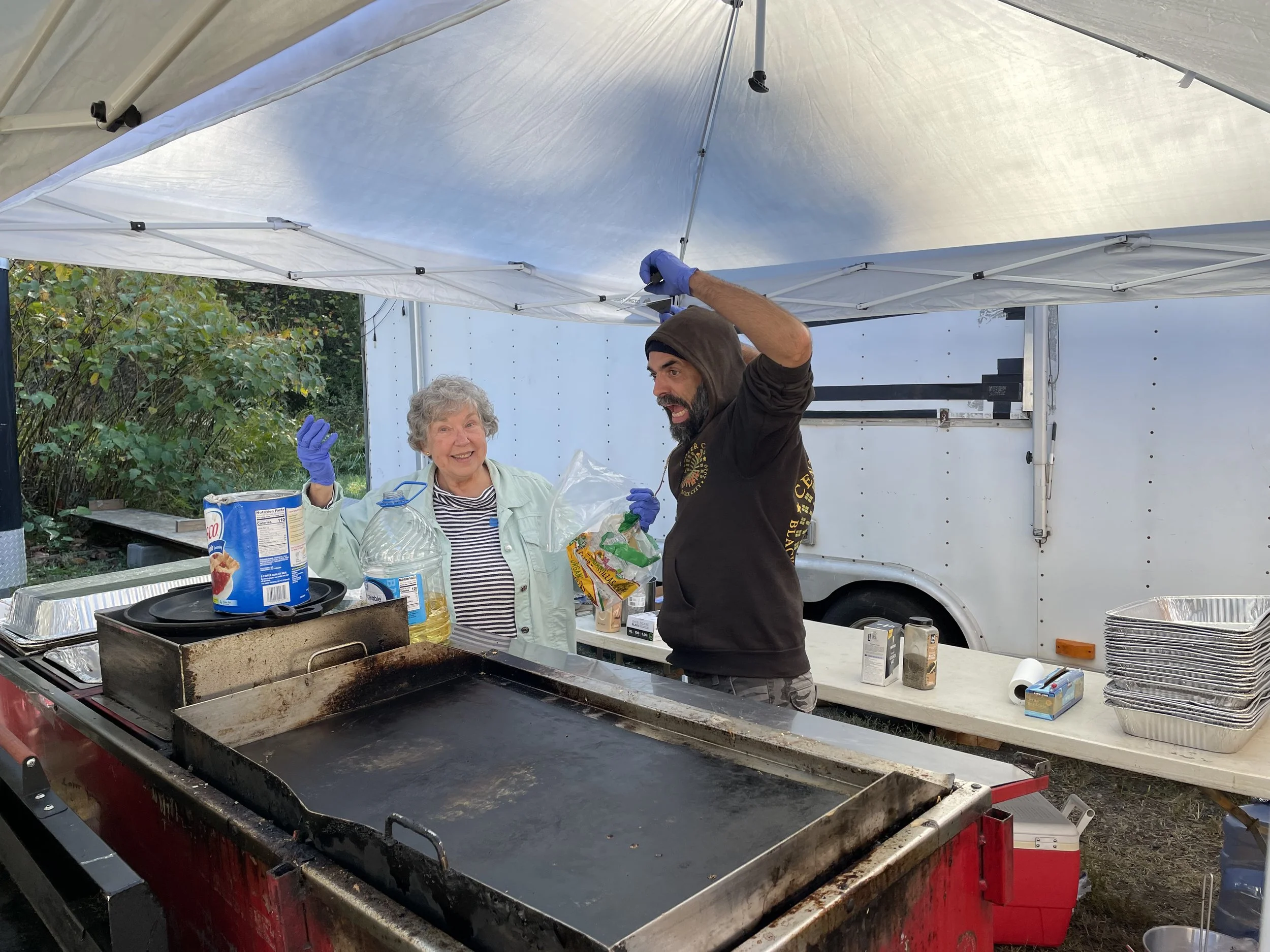 A man and an elderly woman are preparing food or cooking outdoors under a white canopy tent. The man, wearing a hoodie, is reaching up and smiling, while the woman, with curly gray hair, is smiling and holding a plastic bag. There is a grill or cooki