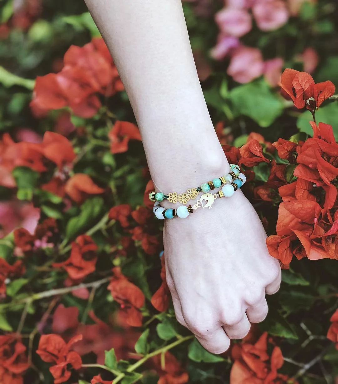 A person's hand and wrist with a beaded bracelet, surrounded by red and pink bougainvillea flowers.