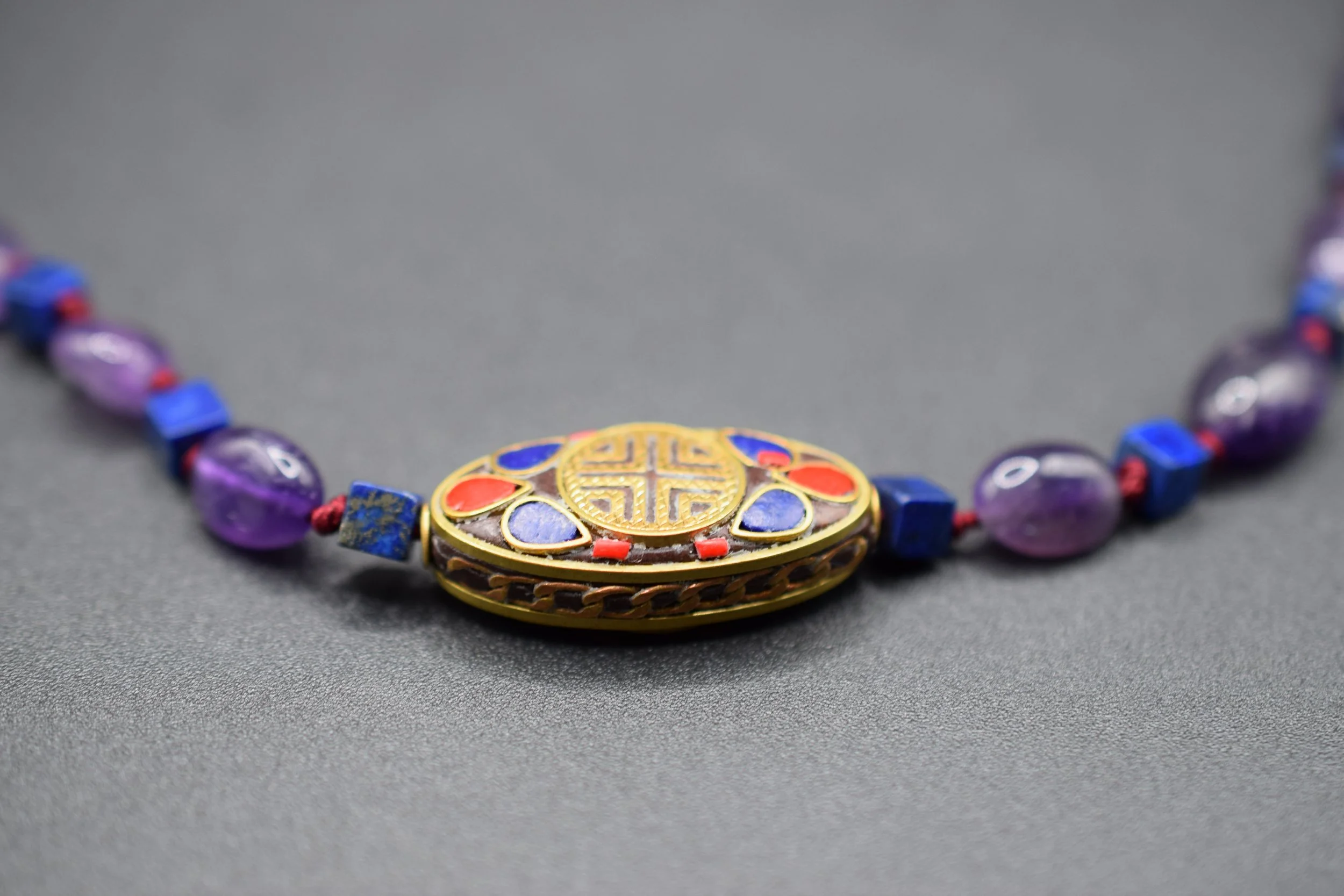 A striking blend of mysticism and elegance, this handcrafted necklace features deep purple amethyst beads paired with vivid blue lapis lazuli cubes, each knotted with crimson thread for added depth and contrast. Accented with intricately detailed inl