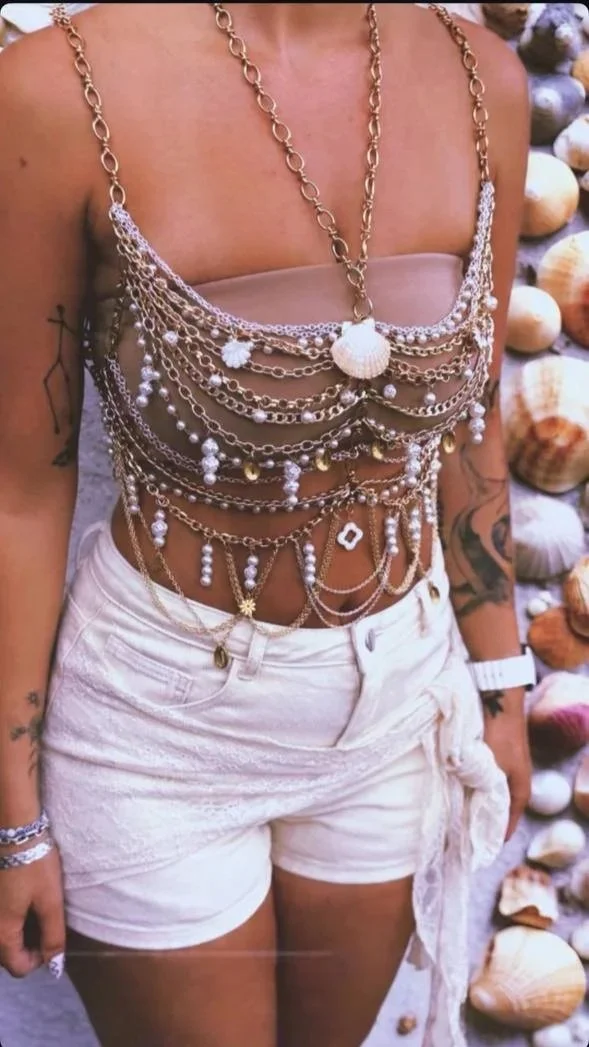 A woman wears multiple layered gold and pearl chains as a top, paired with white shorts and stands against a backdrop of seashells.