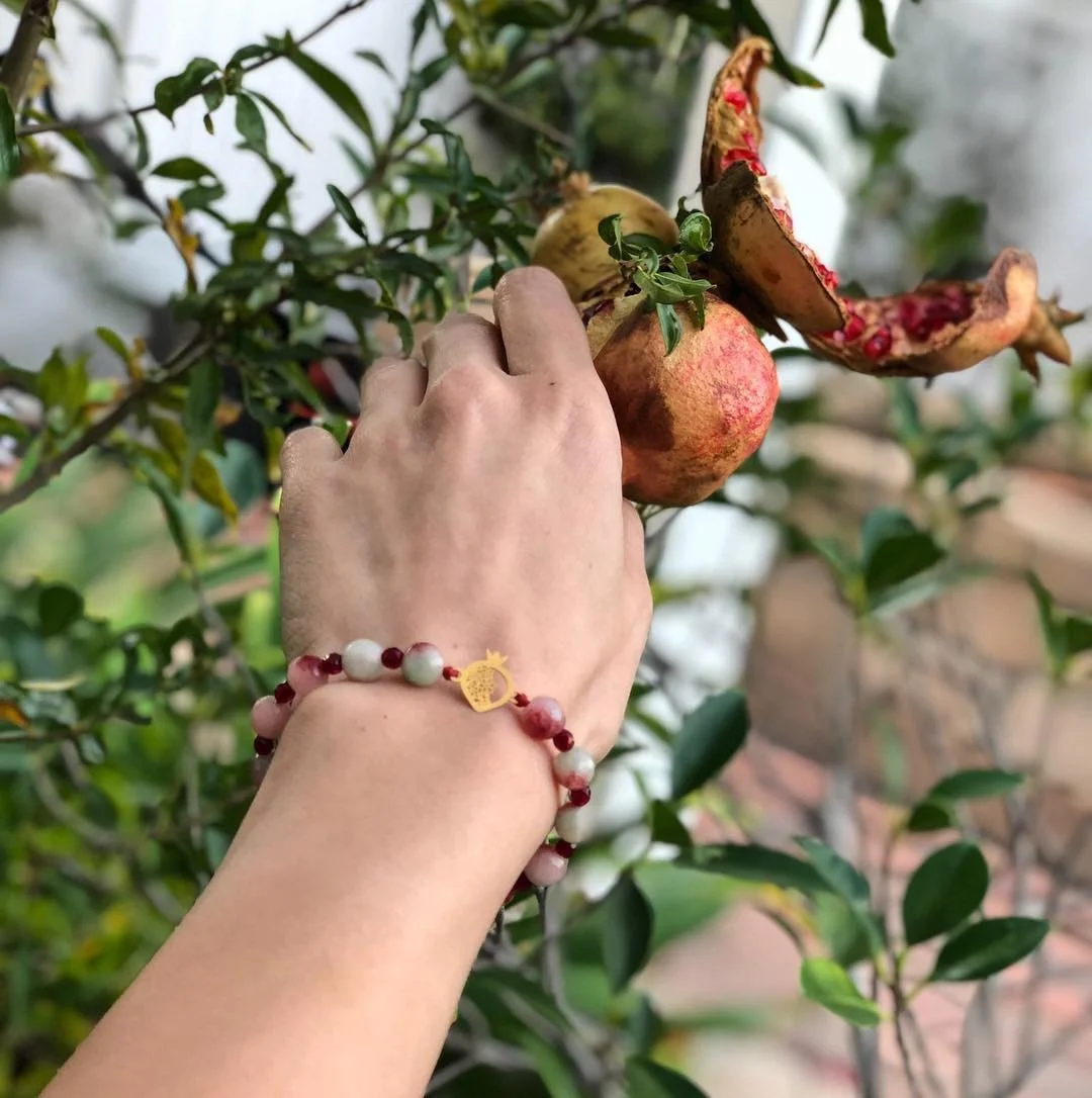 Hand reaching for a pomegranate on a tree, with some fruit split open revealing red seeds, surrounded by green leaves.