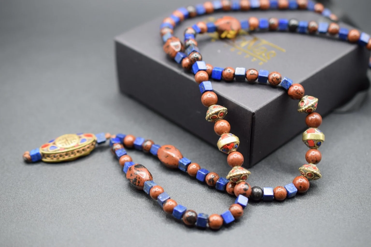 A beaded necklace featuring copper, blue, black, and gold beads, with a pendant, resting on a black surface with a black jewelry box in the background.