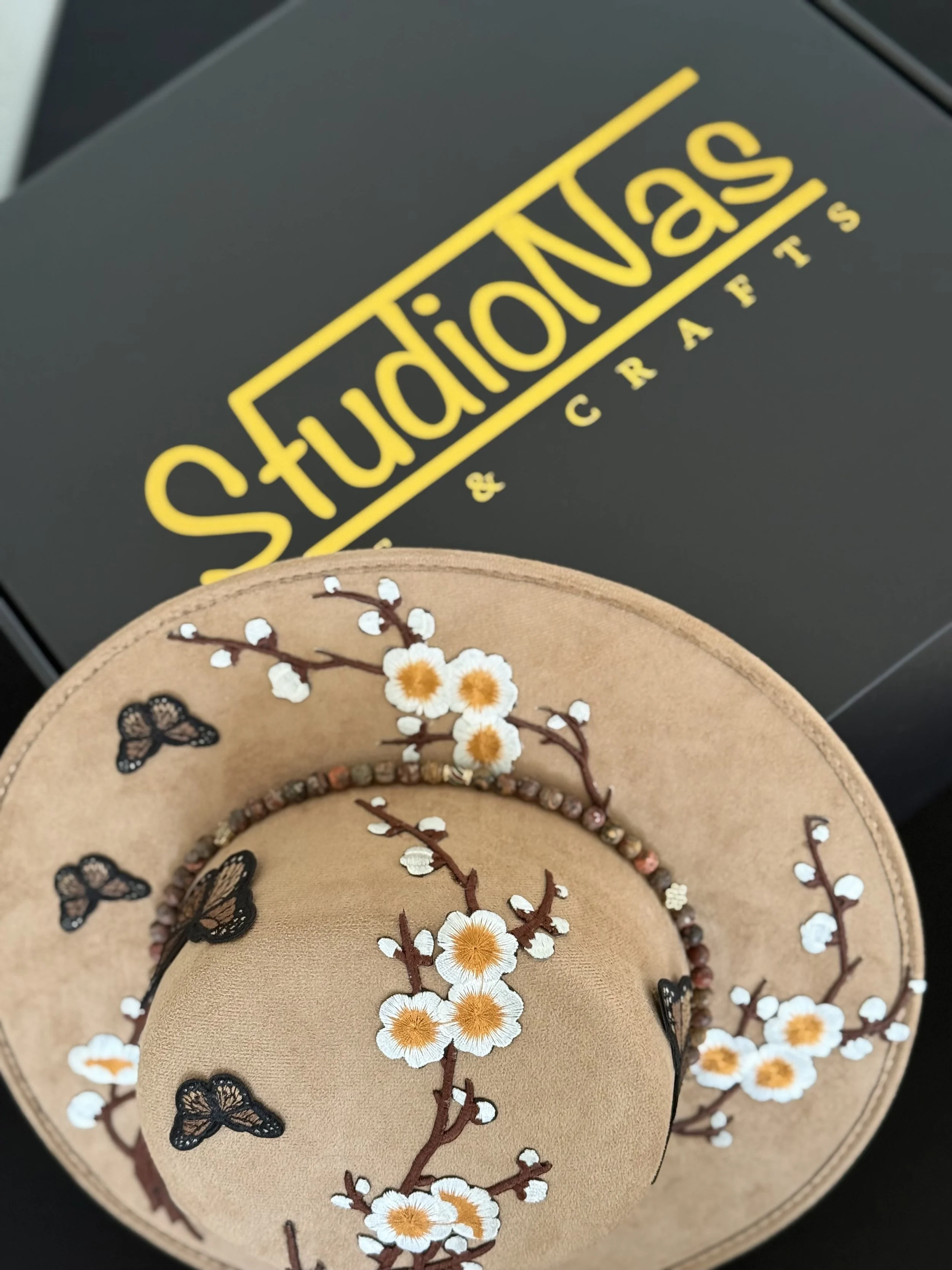 A beige hat with embroidered white and orange flowers, black butterflies, and small beaded accents, placed on top of a black box with yellow lettering reading StickerNast & Crafts.