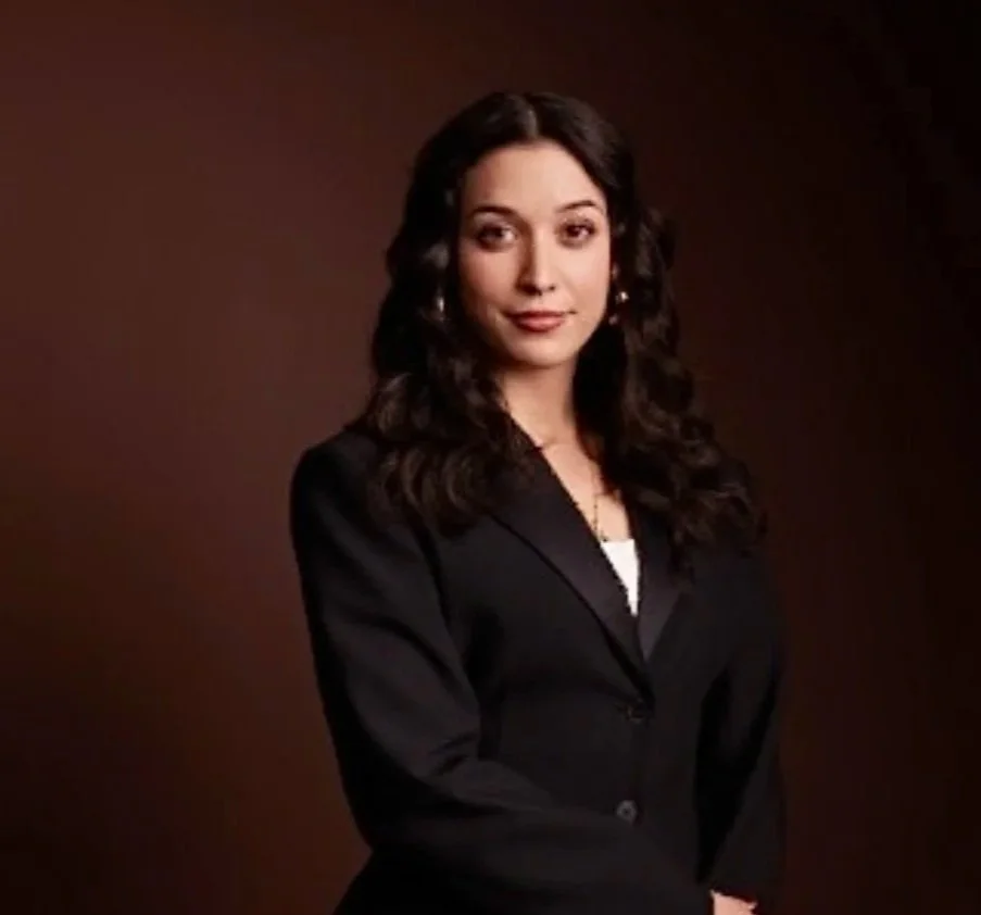 A woman with dark, curly hair wearing a black blazer against a brown background.