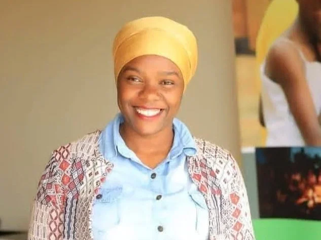 A man smiling outdoors with trees in the background, wearing a plaid shirt.
