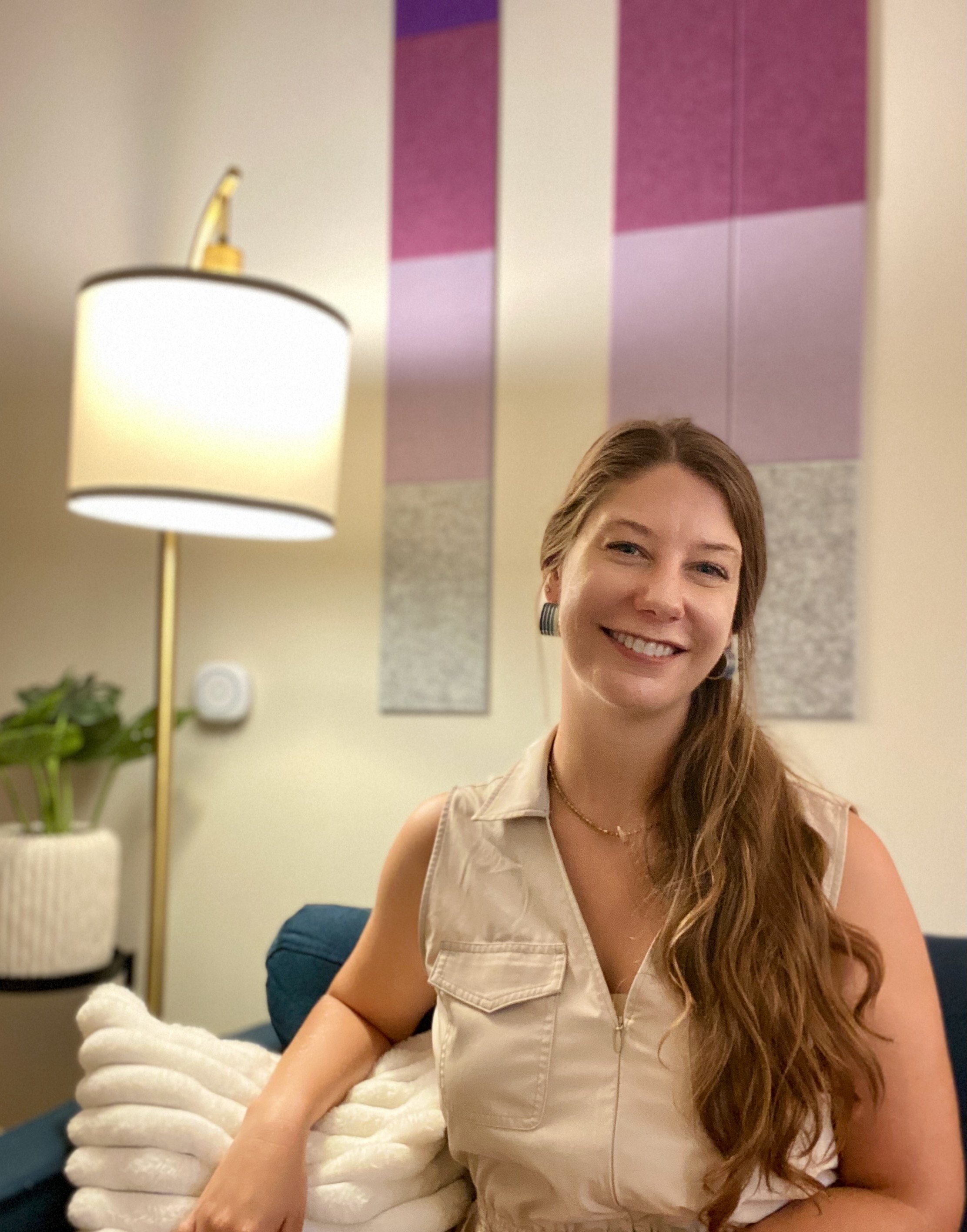 A smiling woman with long, wavy hair sitting on a couch in a living room, with a decorative wall art behind her and a lamp on a stand to her left.