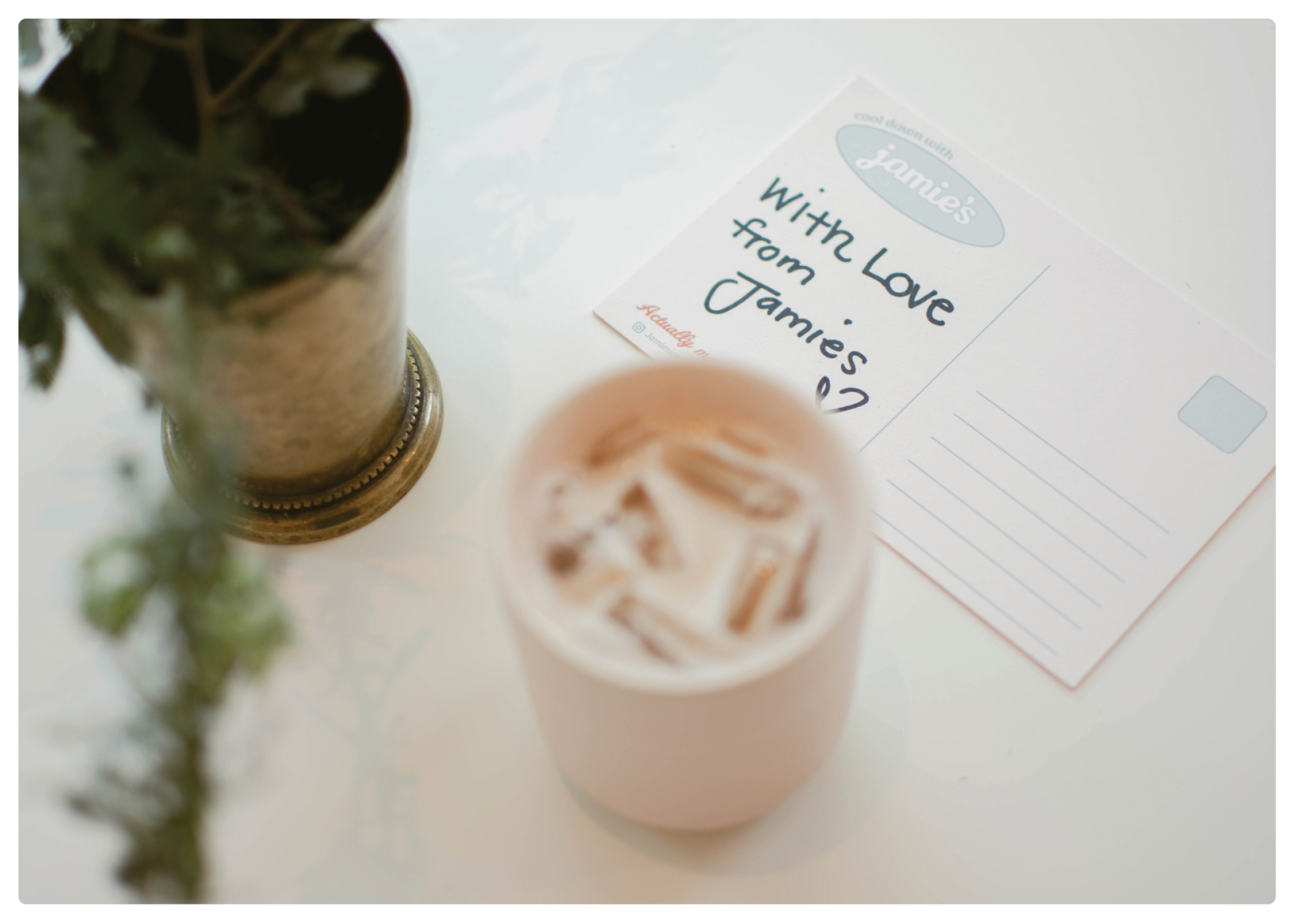A white table with a potted plant, a glass of iced coffee with ice cubes, and a handwritten greeting card that says 'With Love from Jammie's' with a small heart drawing.
