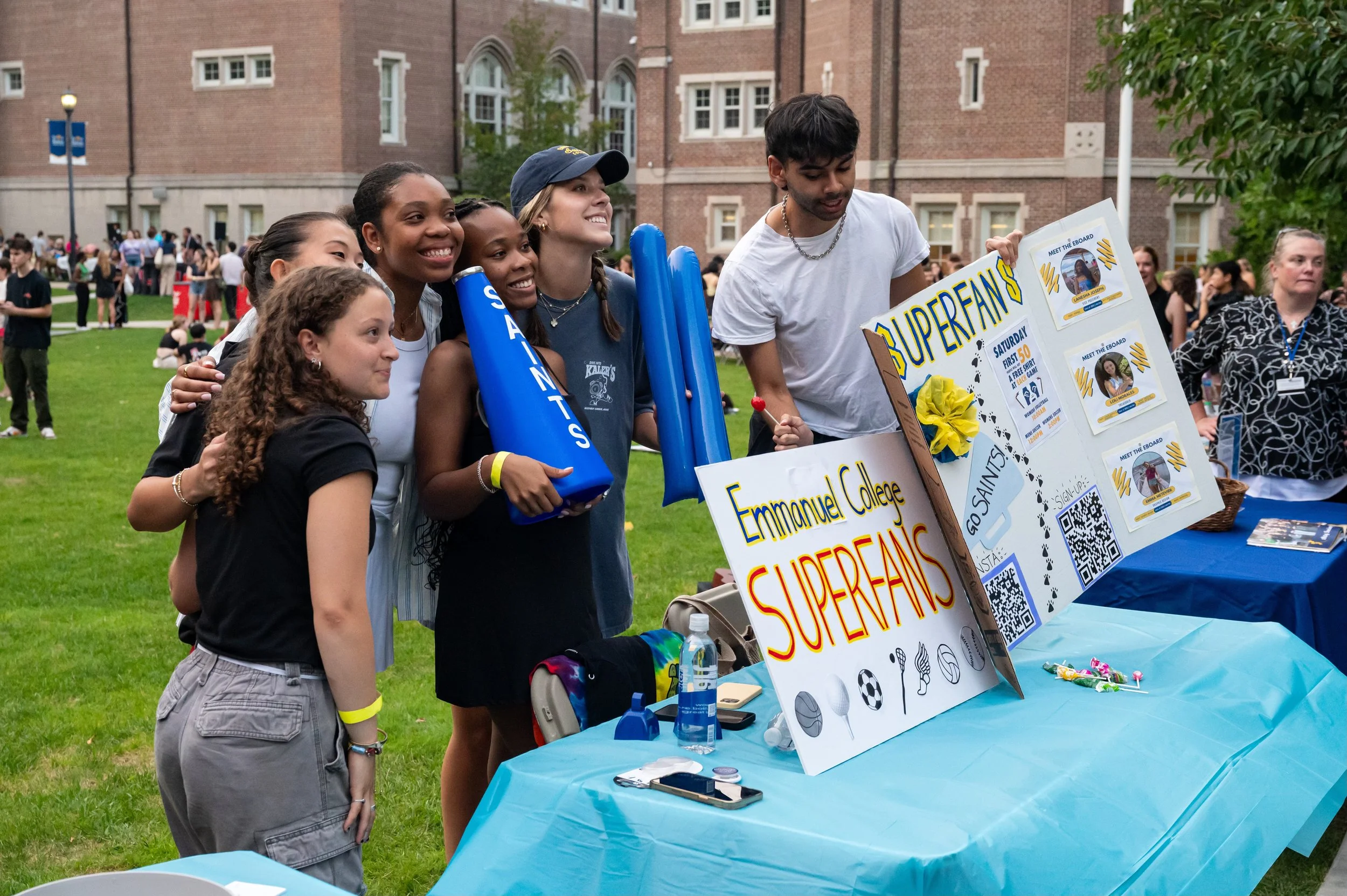 Group of diverse students at Emmanuel College's Superfans booth on a college campus, some smiling and holding balloons, with a table displaying banners, QR codes, and promotional materials for the event.