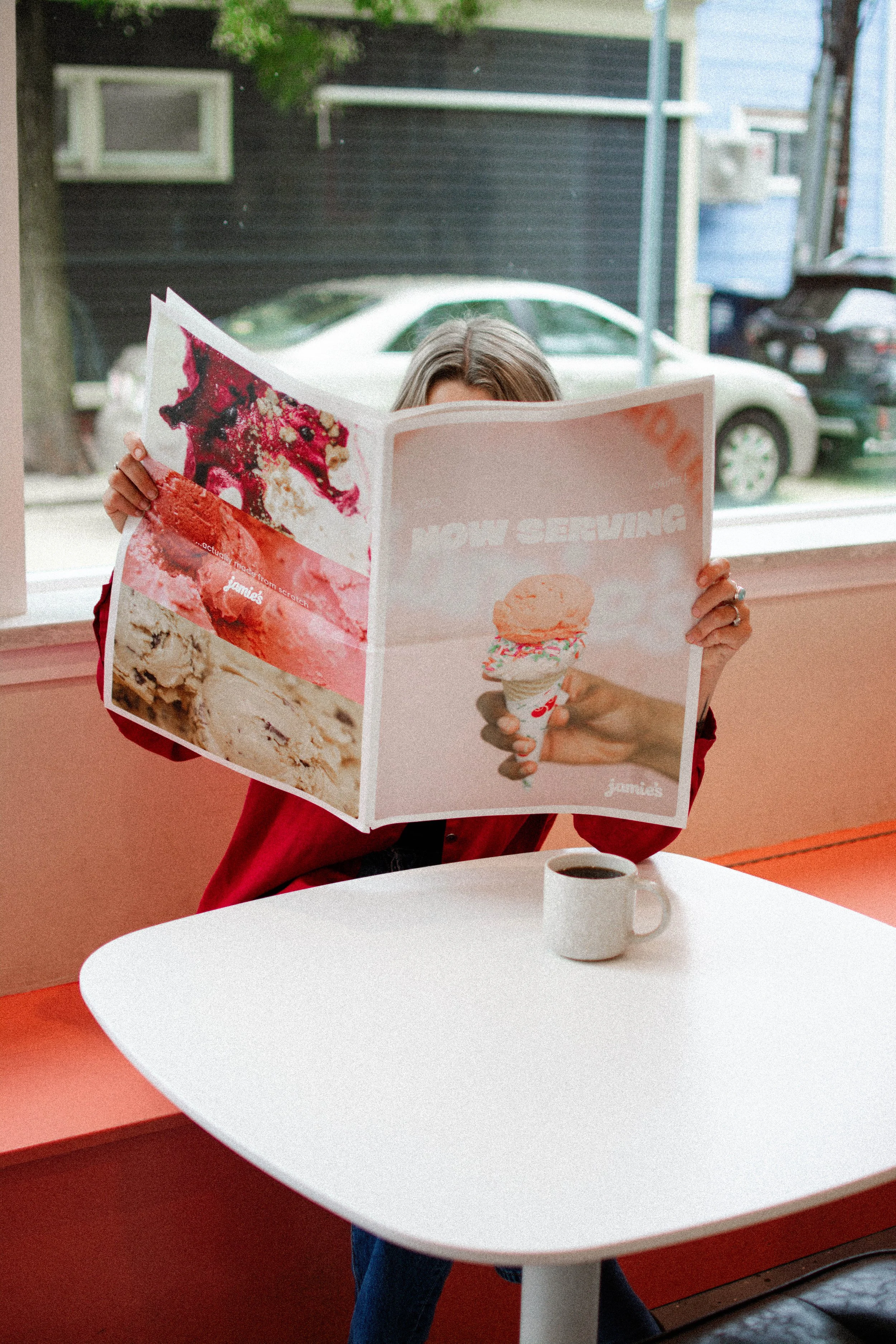 A person sitting at a table reading a large colorful ice cream ad poster inside a cafe, with a white coffee mug on the table and cars visible outside through the window.