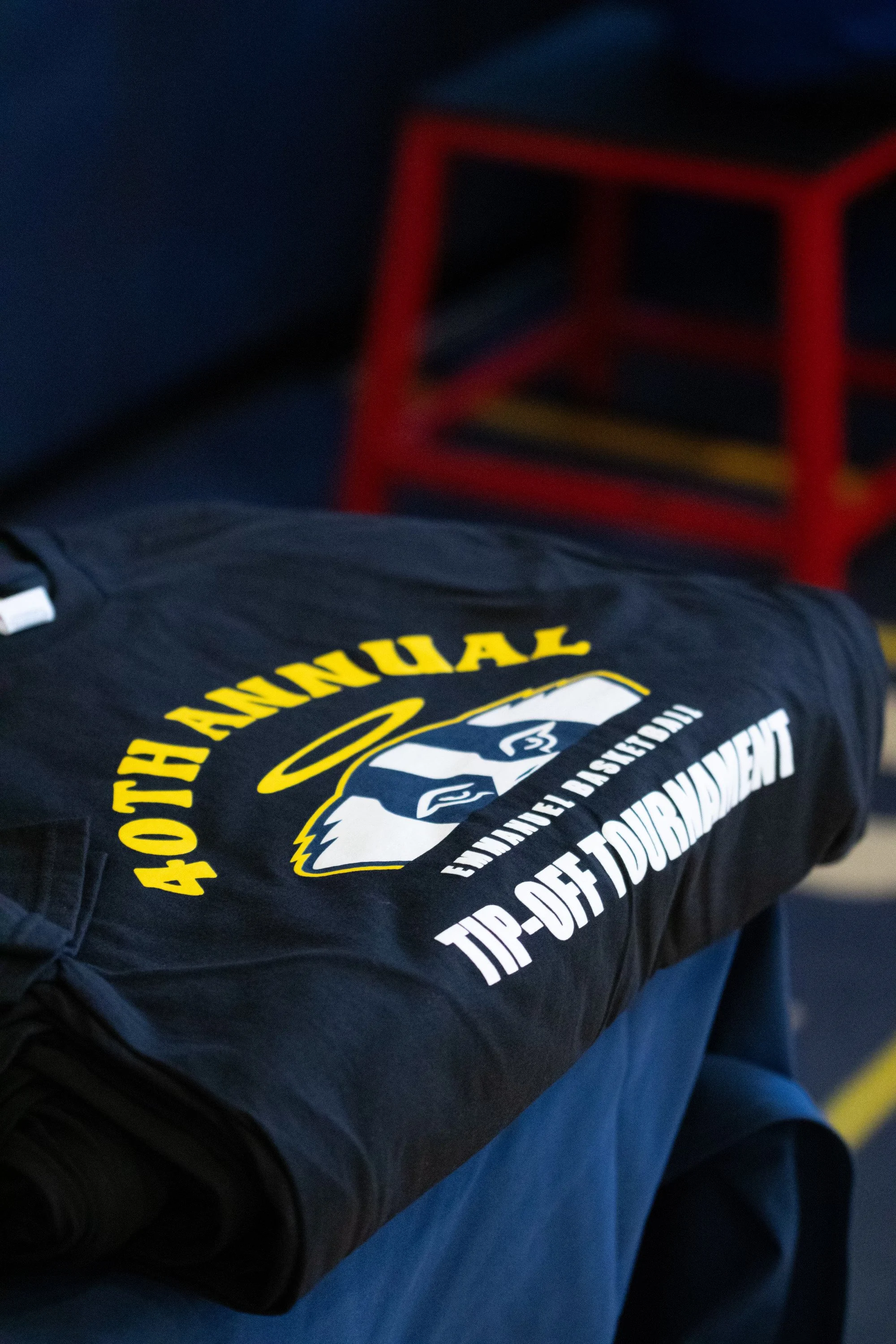 A folded black t-shirt with a logo and text for a sports team, resting on a blue surface, with a red chair in the background.