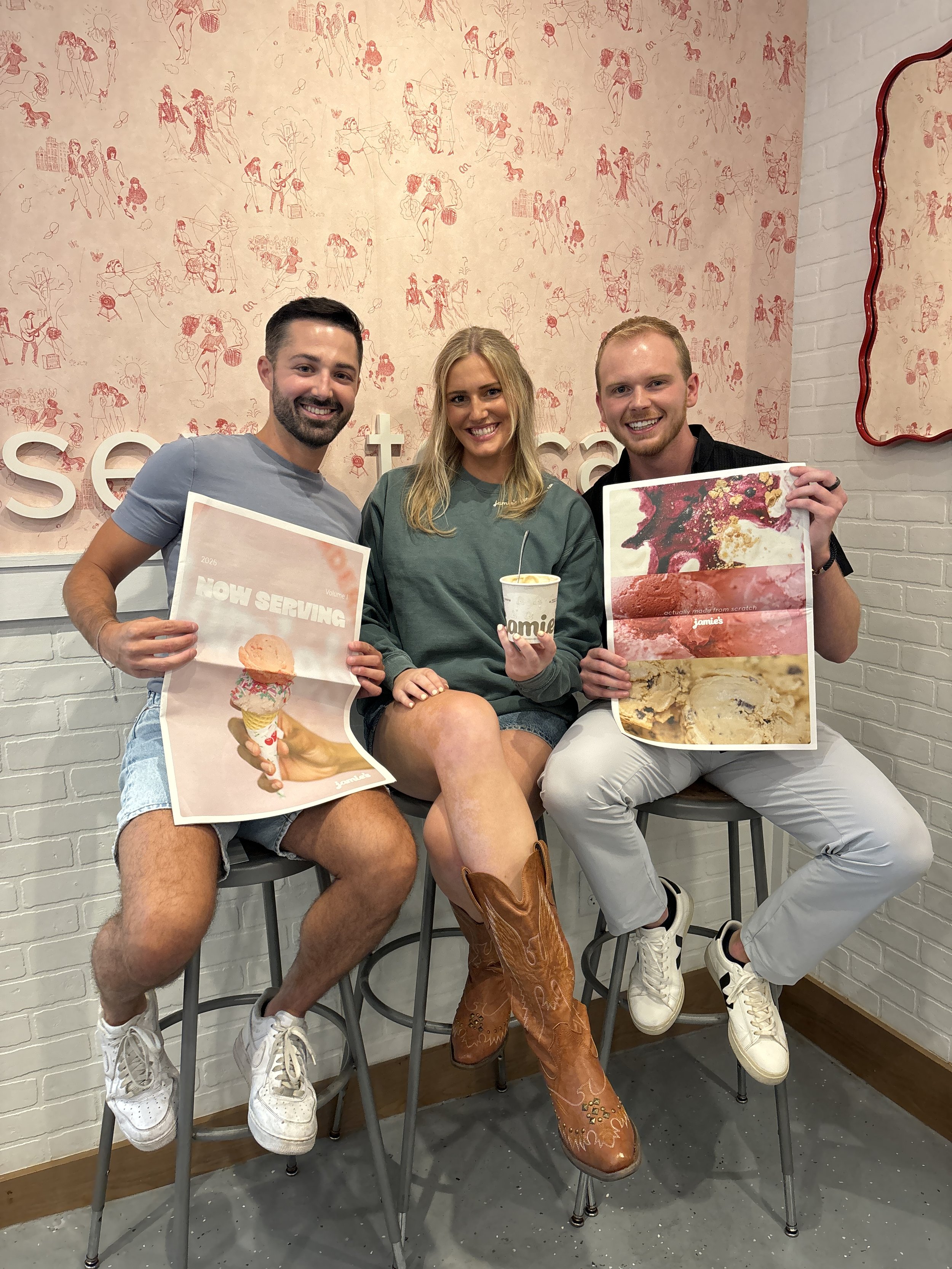 Three people sitting on bar stools against a wall holding promotional posters for ice cream. The person in the middle, a woman, is holding a cup of ice cream. The man on the left is holding a poster with an ice cream cone and the words 'Now Serving'. The man on the right is holding a poster showing different ice cream flavors. They are smiling, and the woman is wearing cowboy boots.