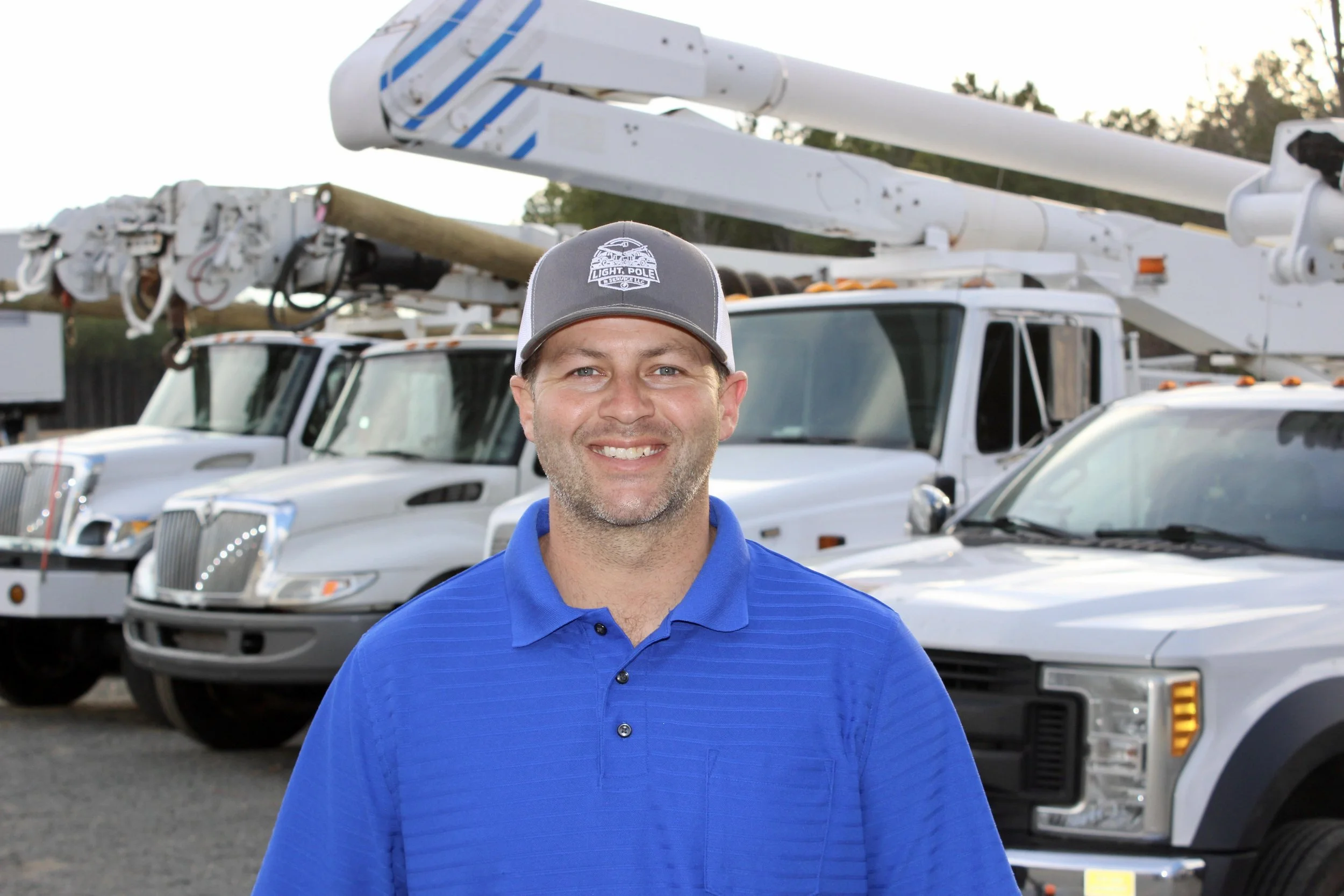 A smiling man in a blue polo shirt and gray cap standing in front of a row of utility trucks with bucket lifts.