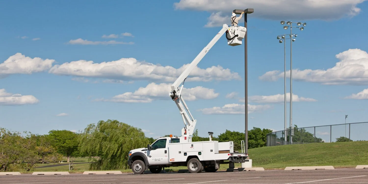 A utility truck with a worker in an aerial lift adjusting a light pole in an outdoor park or sports field setting.