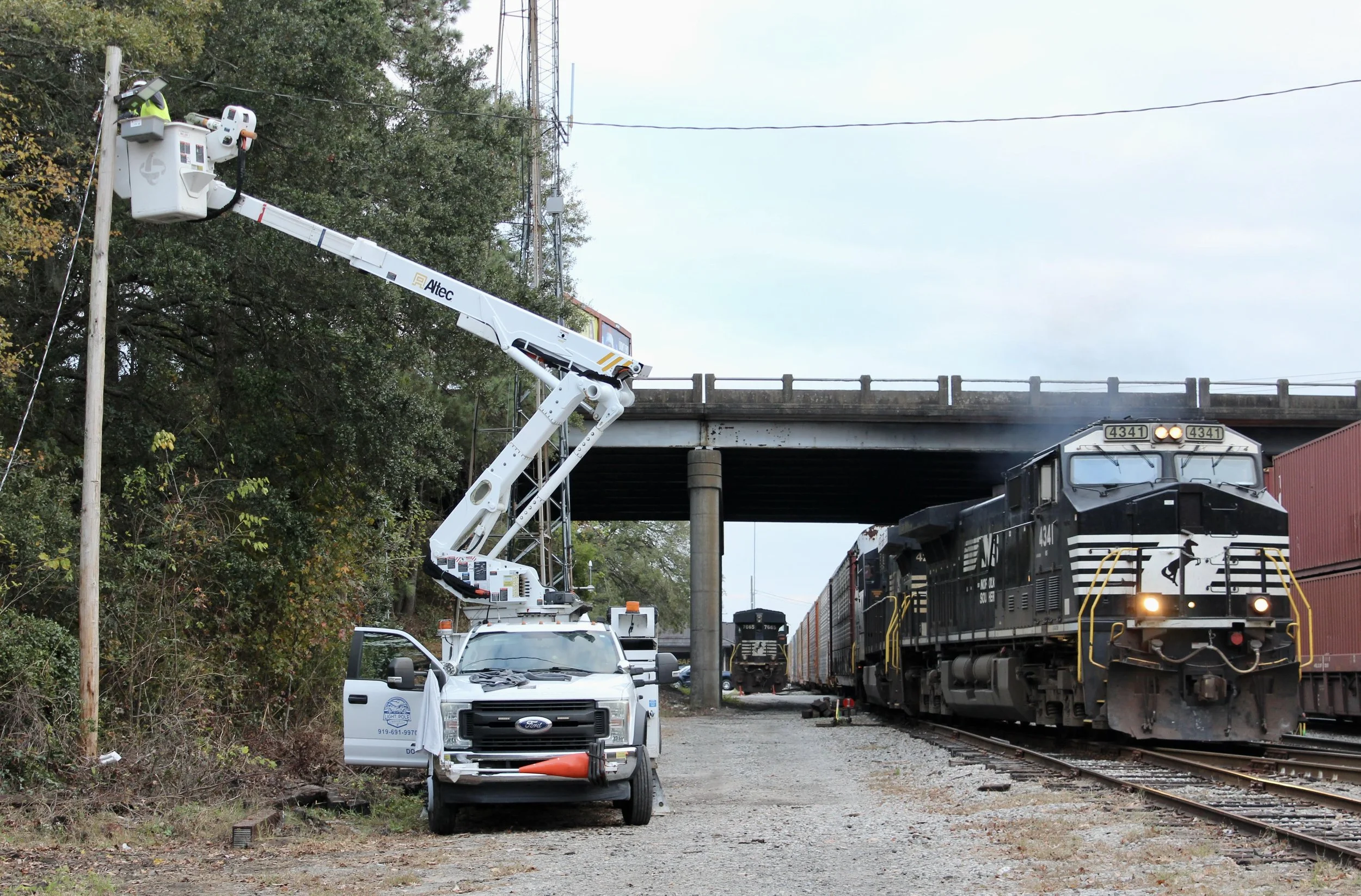 A LPS worker in a bucket lift working on a power pole near a railroad underpass. There are two black locomotives with a number 4341, pulling freight train cars on the tracks.