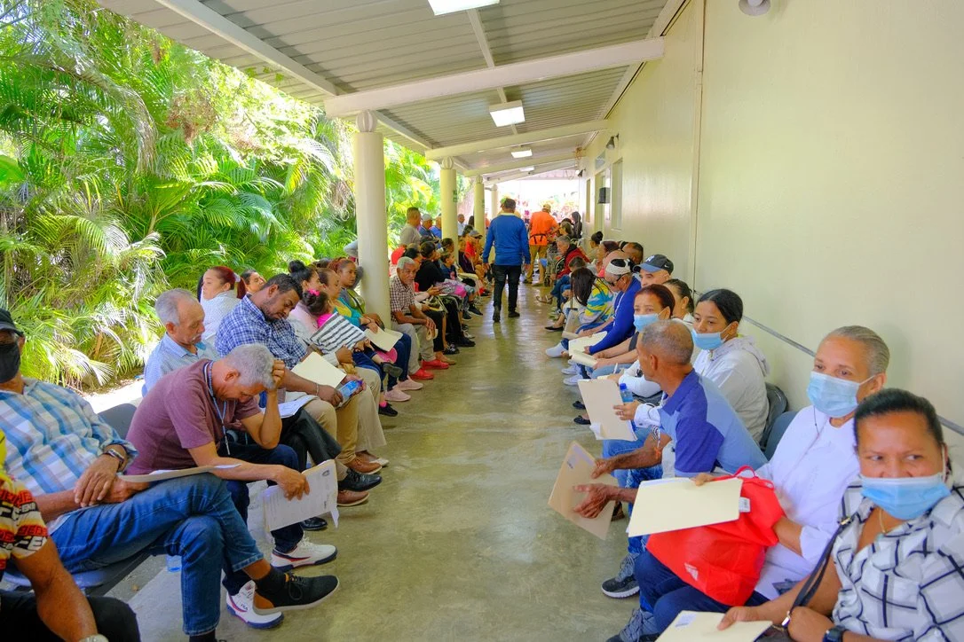 People seated in a long corridor waiting with papers in their hands, some wearing masks, surrounded by greenery outside.