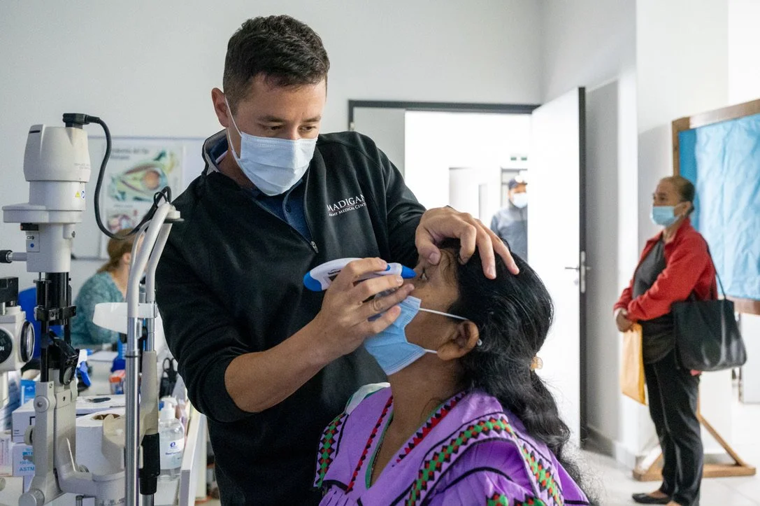 A medical professional performs an eye exam on a woman wearing a colorful outfit, using a handheld device, while both are wearing masks in a clinic setting.