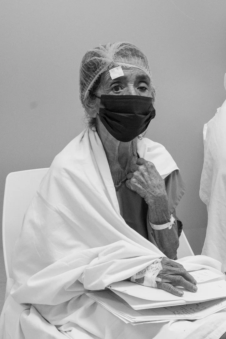 An elderly person wearing a face mask and hair cap sits in a chair. They are covered with a white blanket and have medical papers and a wristband, suggesting a medical setting.