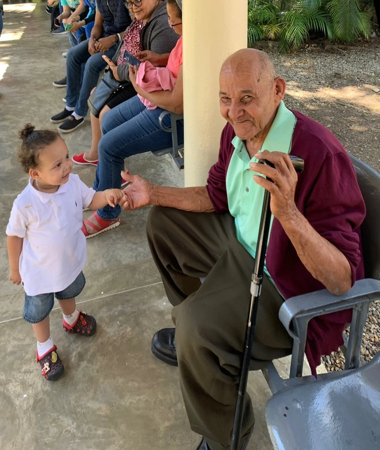 A toddler holding hands with an elderly man sitting on a bench. The man is smiling and holding a cane. There are other people seated in the background.