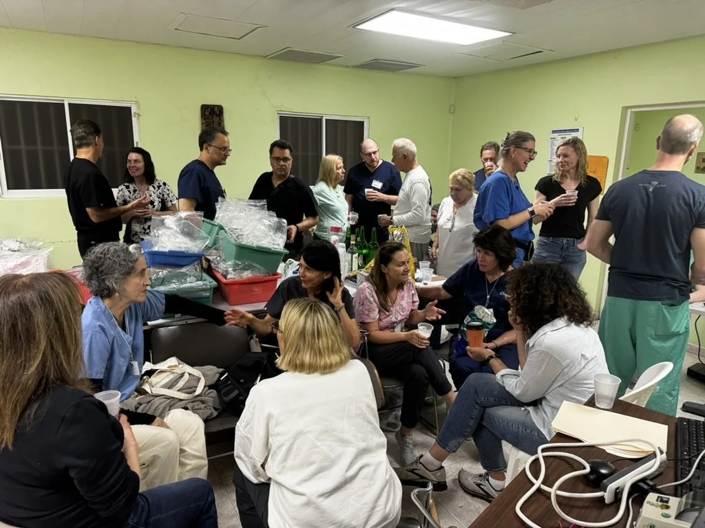 Group of people socializing in a room with green walls, some sitting and some standing, with medical equipment and trays in the background.