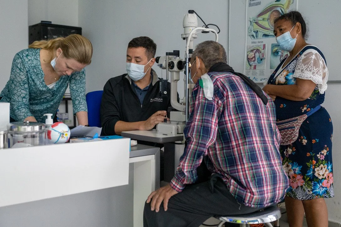 A group of people in a medical office with eye examination equipment. One person is looking into a slit lamp while others, wearing face masks, observe.