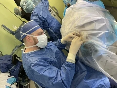 Surgeons in an operating room performing a medical procedure, dressed in blue surgical gowns and gloves, with one surgeon focused on the patient covered in sterile drapes.