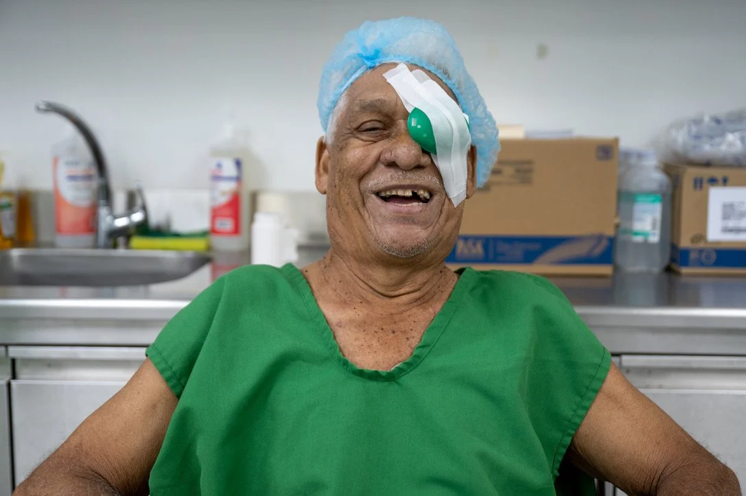 Smiling man in green hospital gown with eye patch and blue hair cap in medical setting.