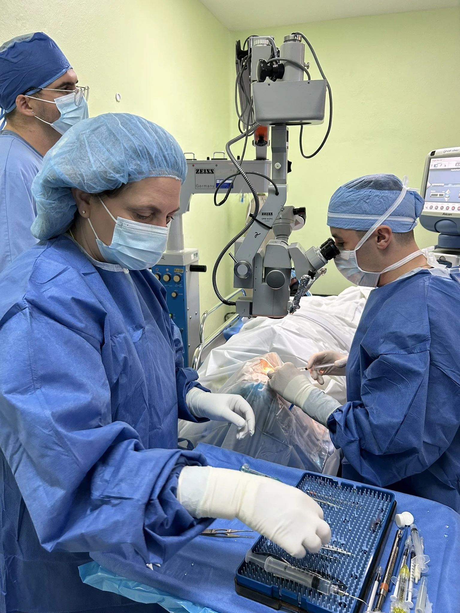 Surgeons in blue surgical gowns and masks performing an eye operation under a microscope in an operating room.