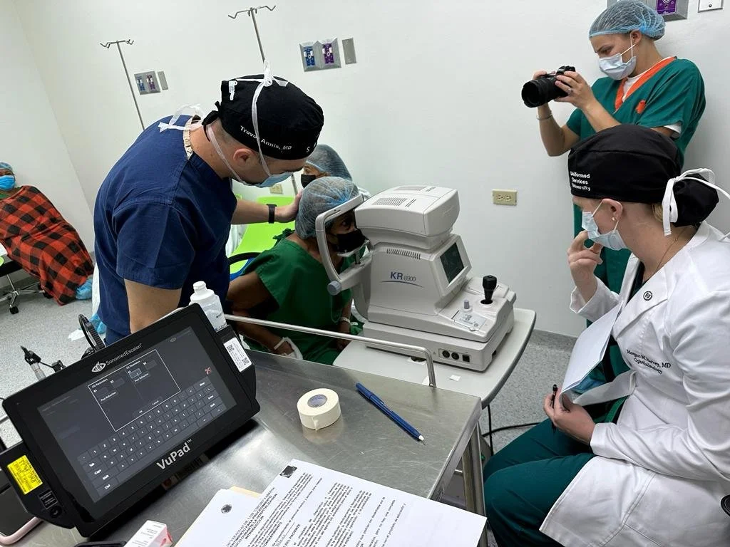 A group of medical professionals in a clinic conducting an eye examination using an optical machine. One person is operating the device while another is observing the results on a tablet. A third person is taking photographs, and there's a patient po