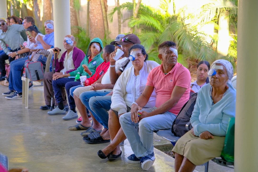 A group of people sitting in a shaded outdoor area, many wearing eye patches, possibly post-eye surgery, with greenery in the background.