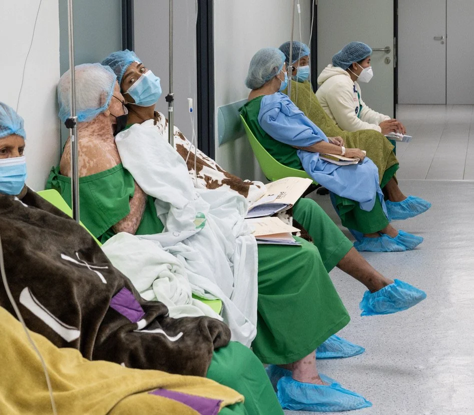 Patients wearing surgical gowns, hair caps, and masks sit in a hospital waiting area with IV drips, covered in blankets.