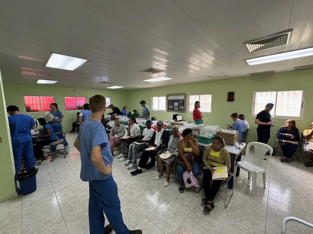 A group of people in a waiting room or clinic with several individuals in blue scrubs assisting or interacting with seated individuals. The room has green walls and fluorescent lighting, with various items and paperwork spread across tables. People a
