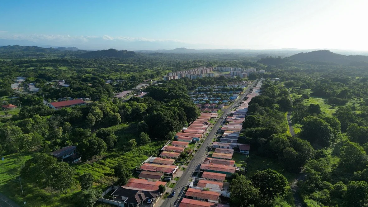 Aerial view of a residential area with rows of houses and tree-covered surroundings, located in a lush green landscape with distant hills and clear blue skies.