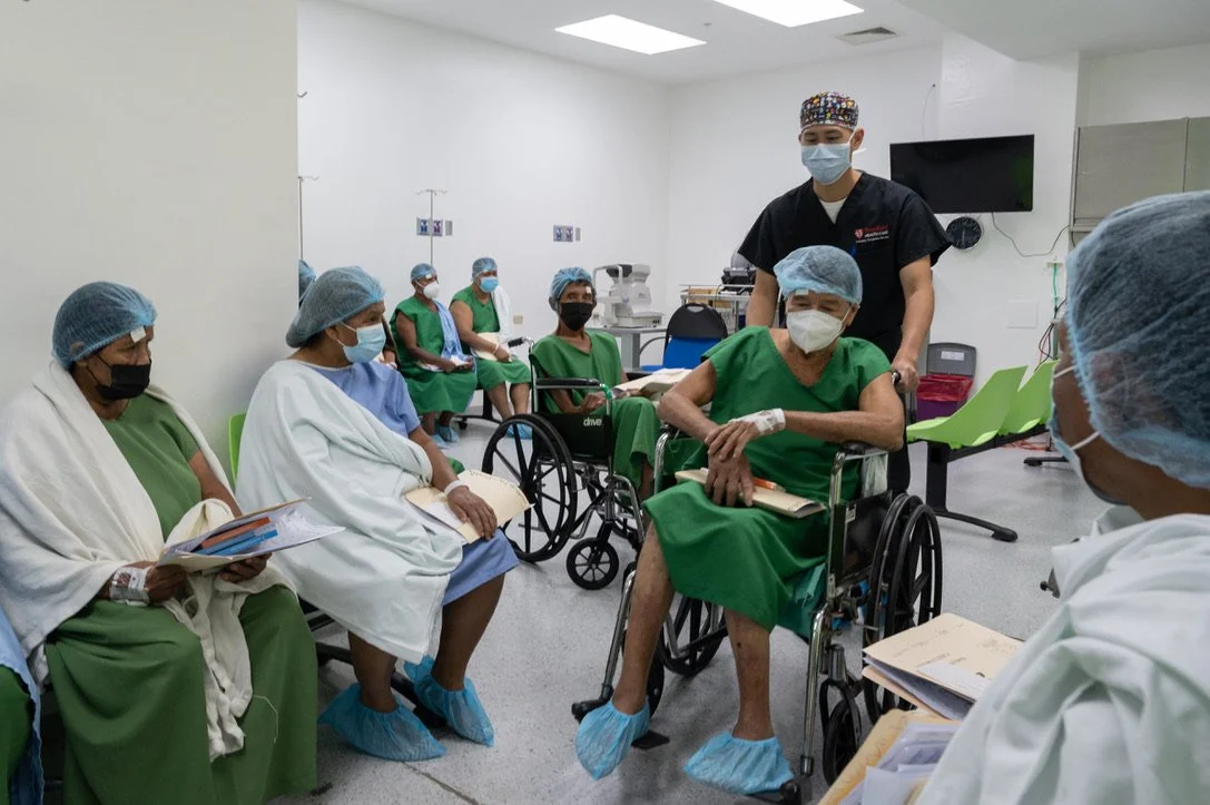 A group of medical patients in hospital gowns and hair caps sit in wheelchairs and chairs in a clinical setting. A healthcare professional in scrubs and a mask stands among them. The patients hold papers and wear face masks and protective footwear.