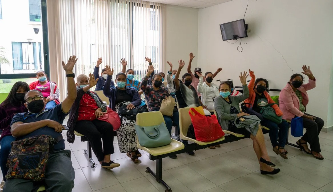 Group of people wearing masks, sitting on chairs in a waiting room, waving to the camera.