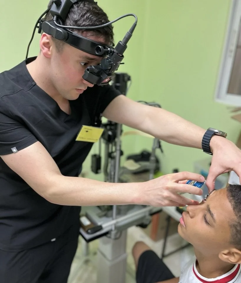 Optometrist performing eye examination on a young boy using specialized equipment and wearing a head-mounted device.