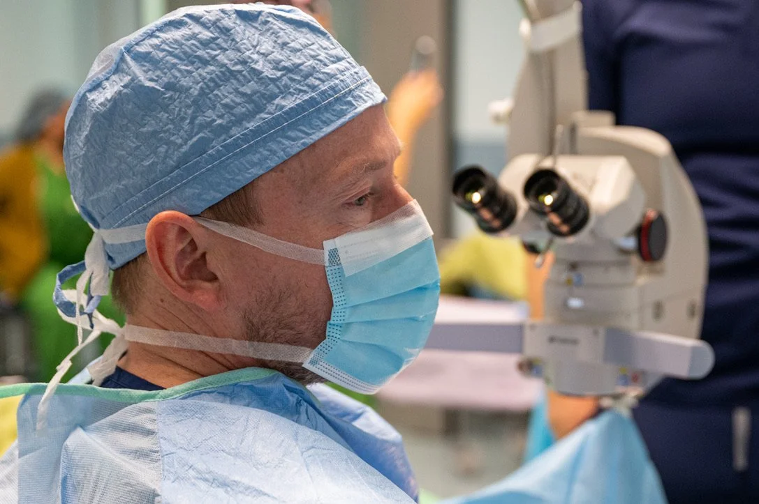 Surgeon in blue surgical attire wearing a surgical mask while using a medical microscope.