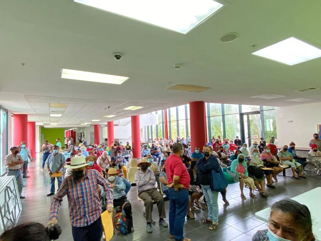 A large group of people wearing face masks sitting and standing in a brightly lit waiting area with red columns and large windows.