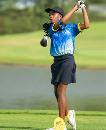Young girl playing golf, holding a golf club, wearing a blue shirt, black shorts, and a cap, standing on a golf course near water.