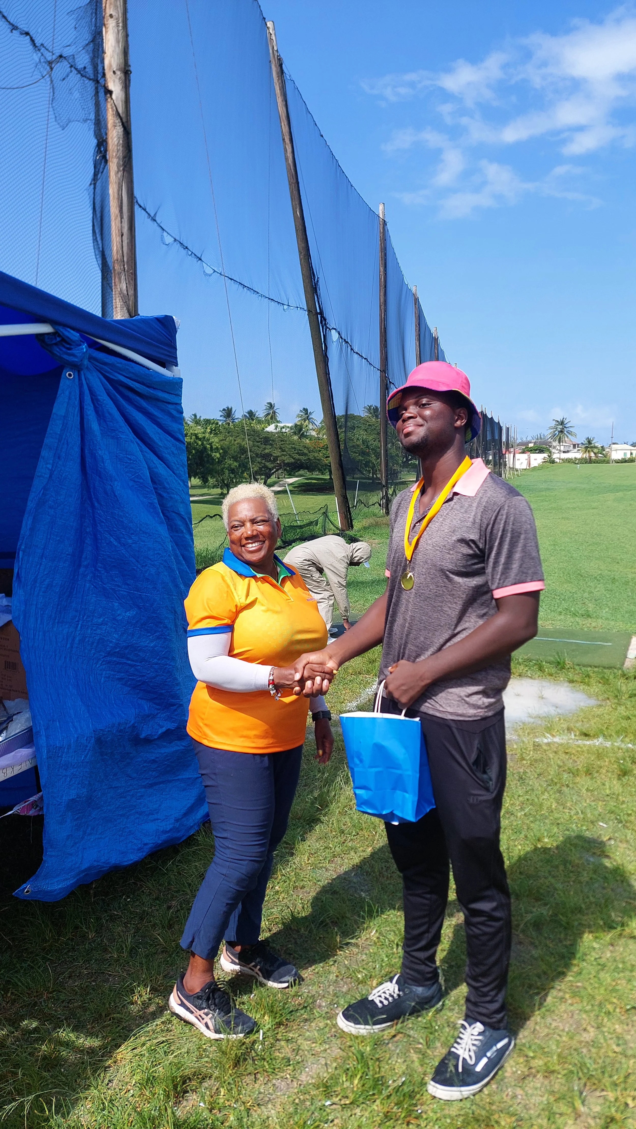 A young man receiving a medal and shaking hands with a smiling woman outdoors on a sunny day. The young man is wearing a pink bucket hat, a gray polo shirt with pink accents, black athletic pants, and black sneakers. He has a blue gift bag and a meda