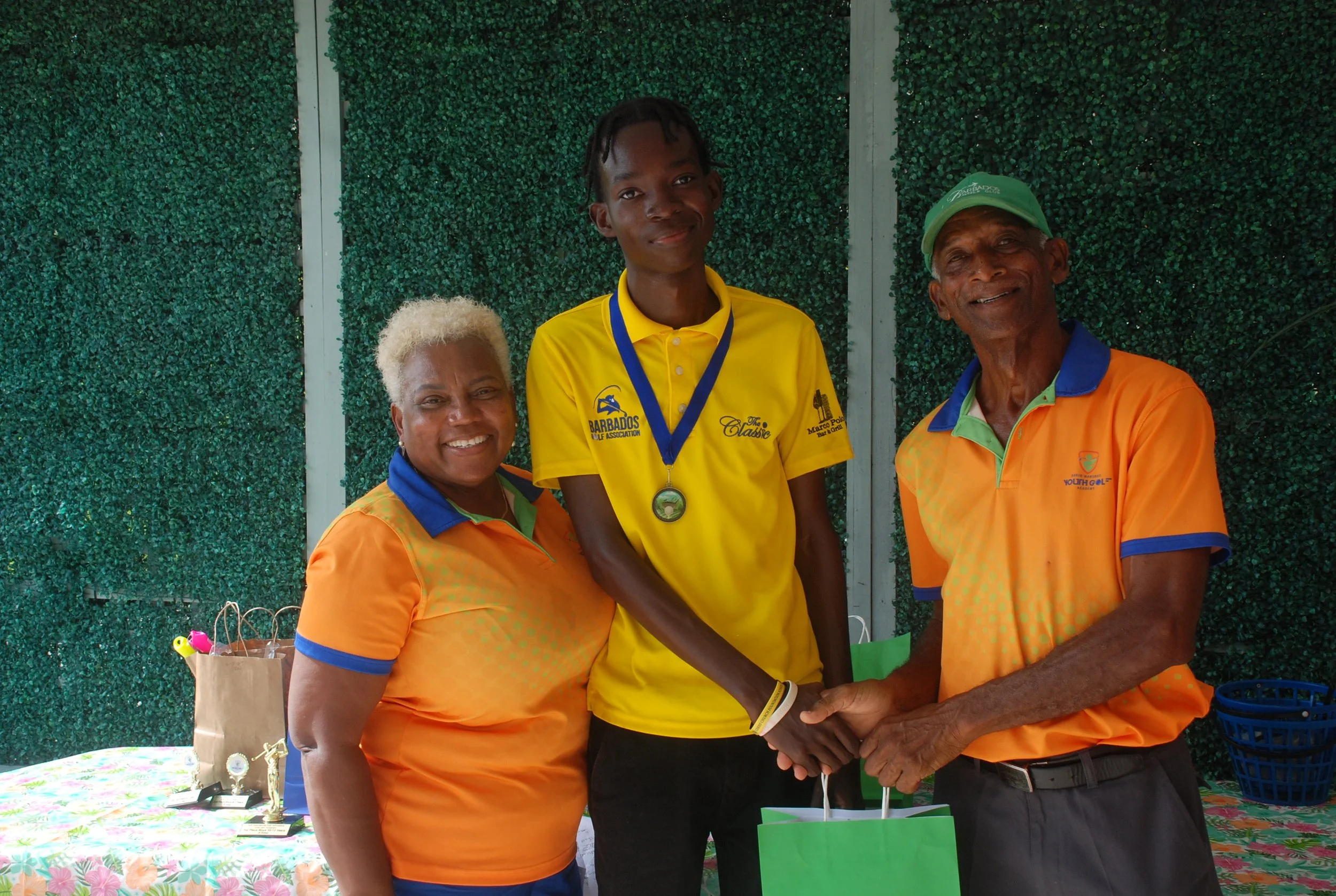 Three people smiling and shaking hands at an outdoor event, with a green hedge background. The young man in the middle is wearing a yellow sports shirt and a medal. The two older individuals on either side are wearing orange shirts with blue collars.