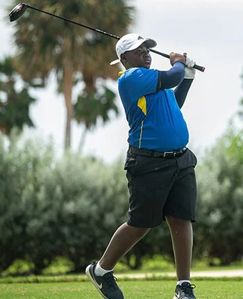 A young boy wearing a white cap, blue shirt, and black shorts, holding a golf club and preparing to swing on a golf course.