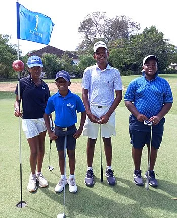 Four children standing on a golf course, dressed in golf clothes, holding golf clubs, with a blue flag and trees in the background.