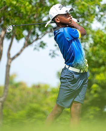 Young boy in a blue shirt and gray shorts playing golf, swinging a club outdoors with trees in the background.