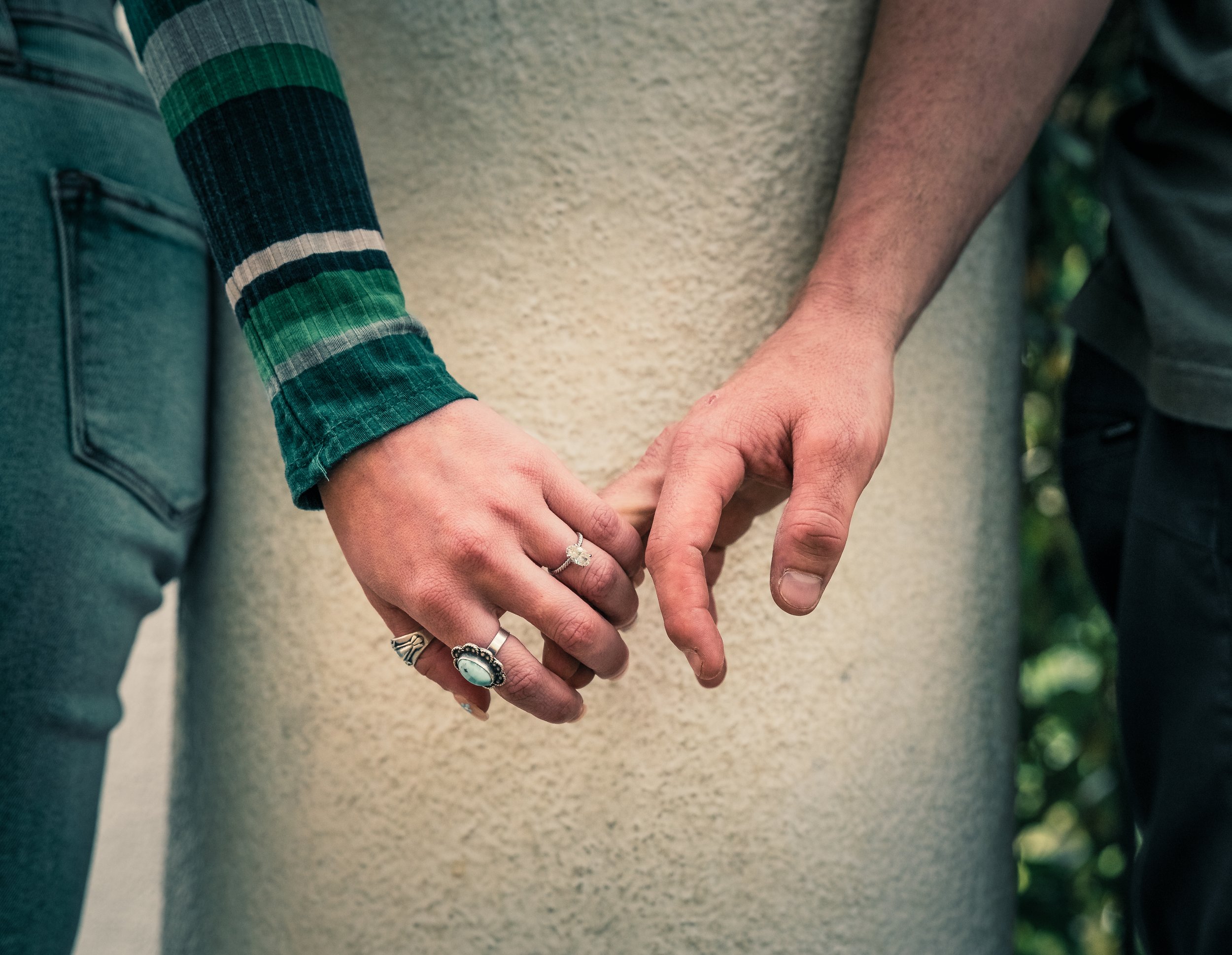 Two people holding hands, with one person wearing multiple rings and a striped green sleeve in an outdoor setting.