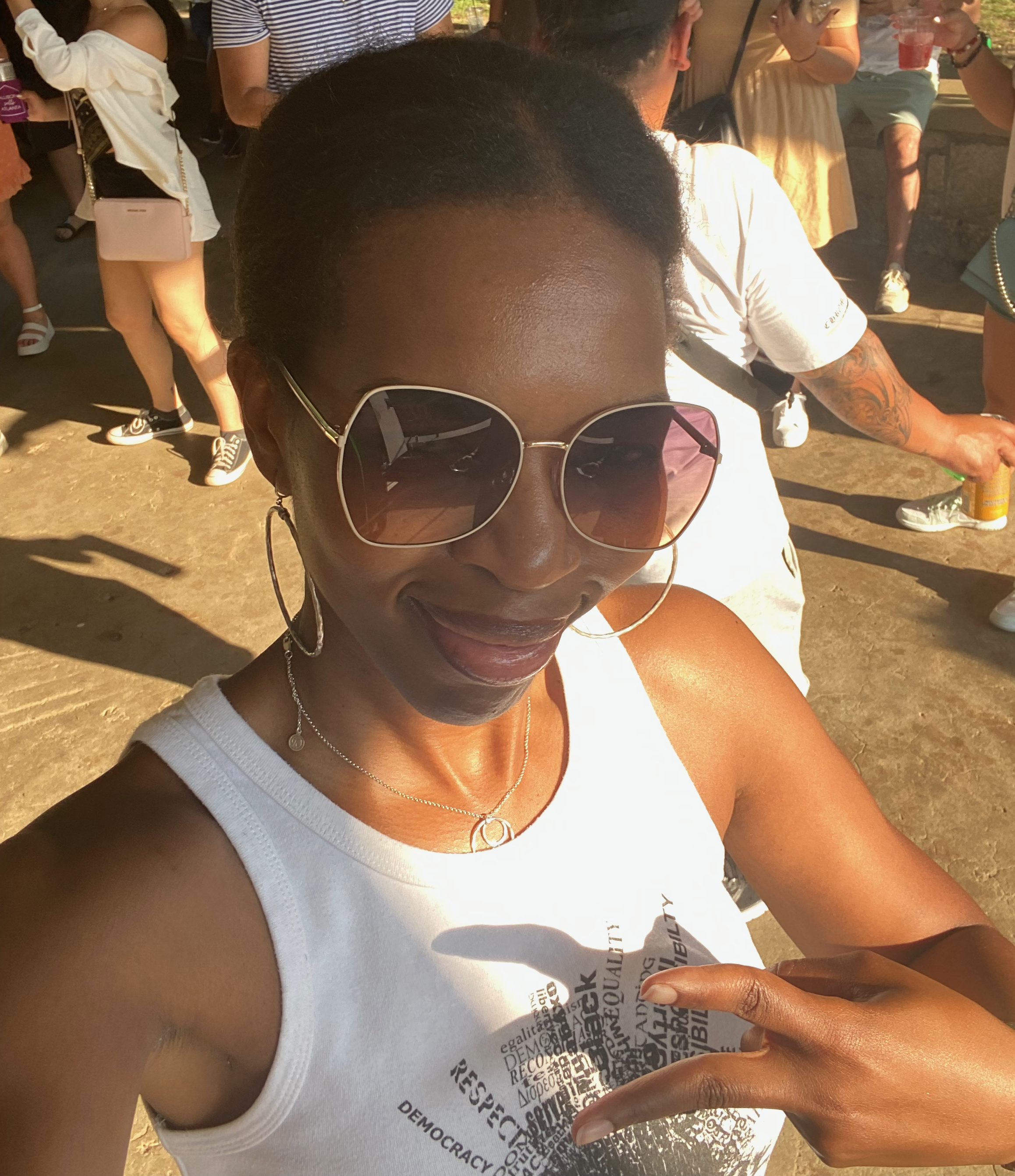A woman wearing sunglasses, a white tank top, and jewelry is smiling. In the background, several people are gathered, some dancing.