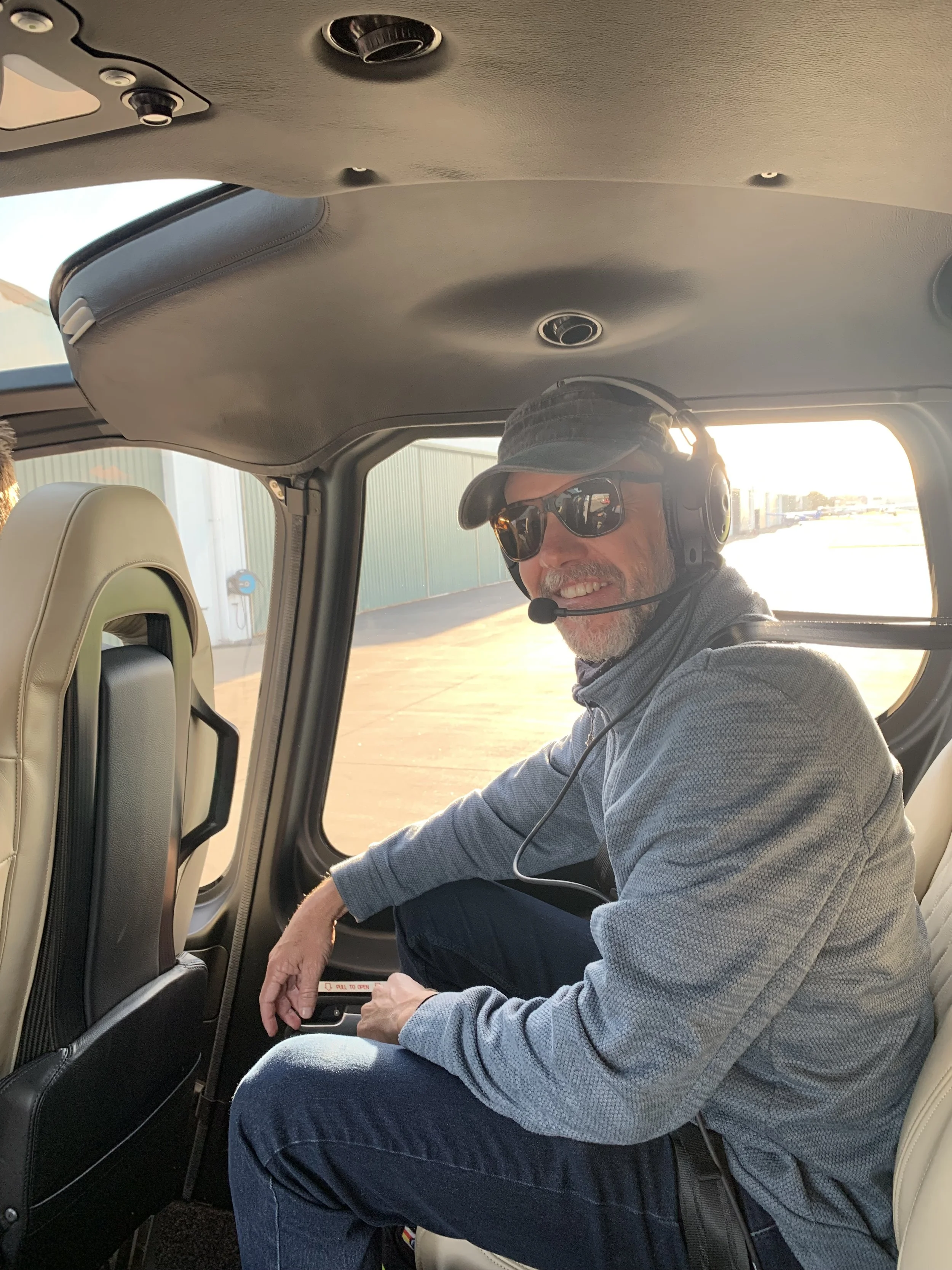 A man sitting in an airplane seat, wearing sunglasses, a camouflage baseball cap, headphones, and a gray sweater, smiling at the camera while holding a small object in his left hand and smiling.