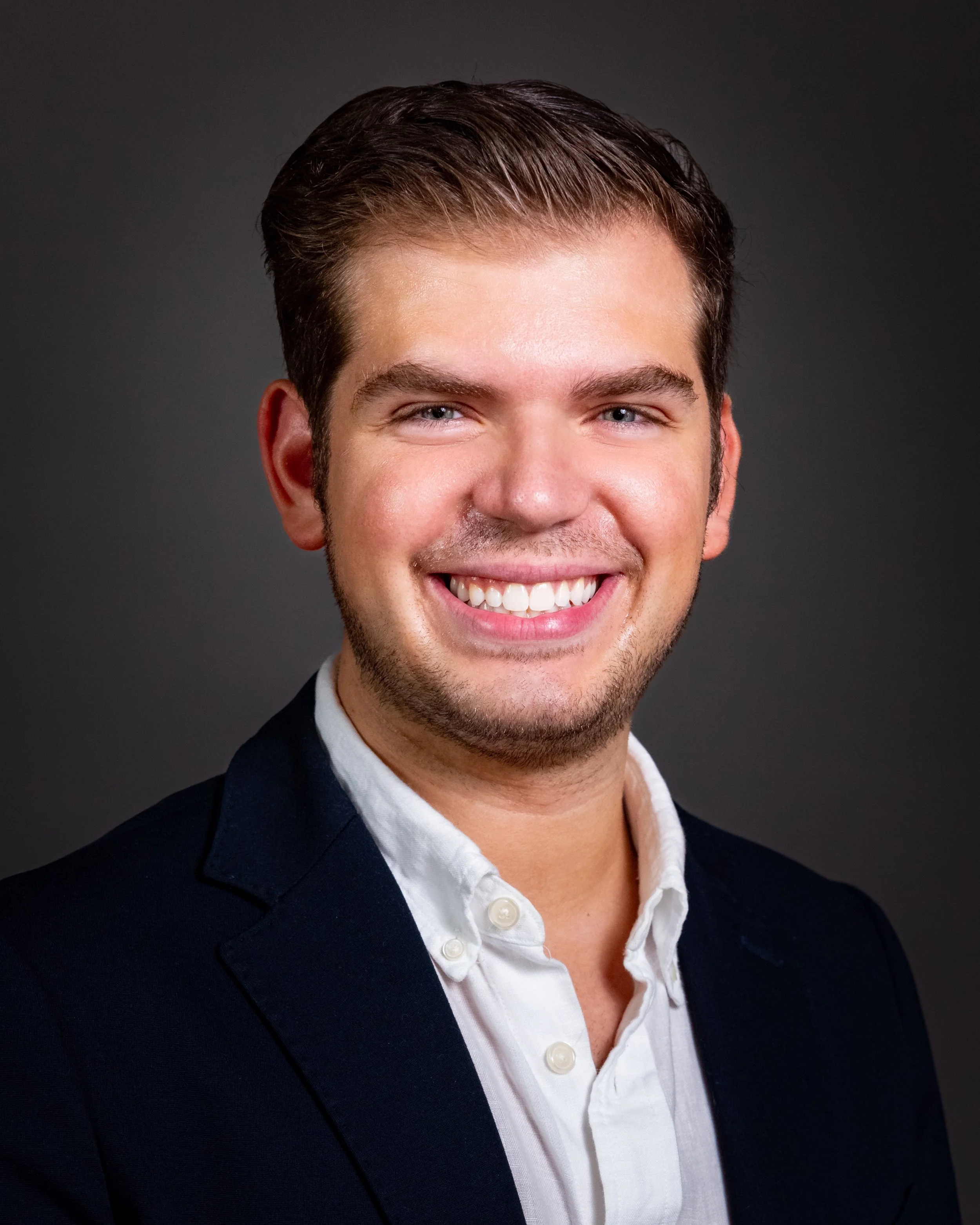 Young man with brown hair, smiling in a suit jacket and white shirt outside in front of historic buildings and a street lamp.
