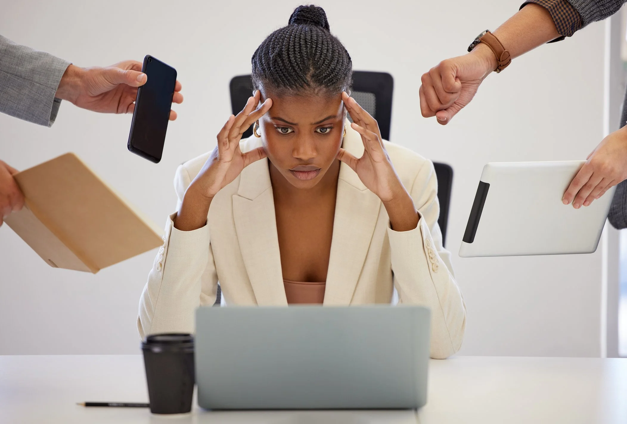 A woman sitting at a desk looking stressed with her hands on her temples, surrounded by multiple hands offering various items like a phone, paper bag, tablet, and coffee cup.