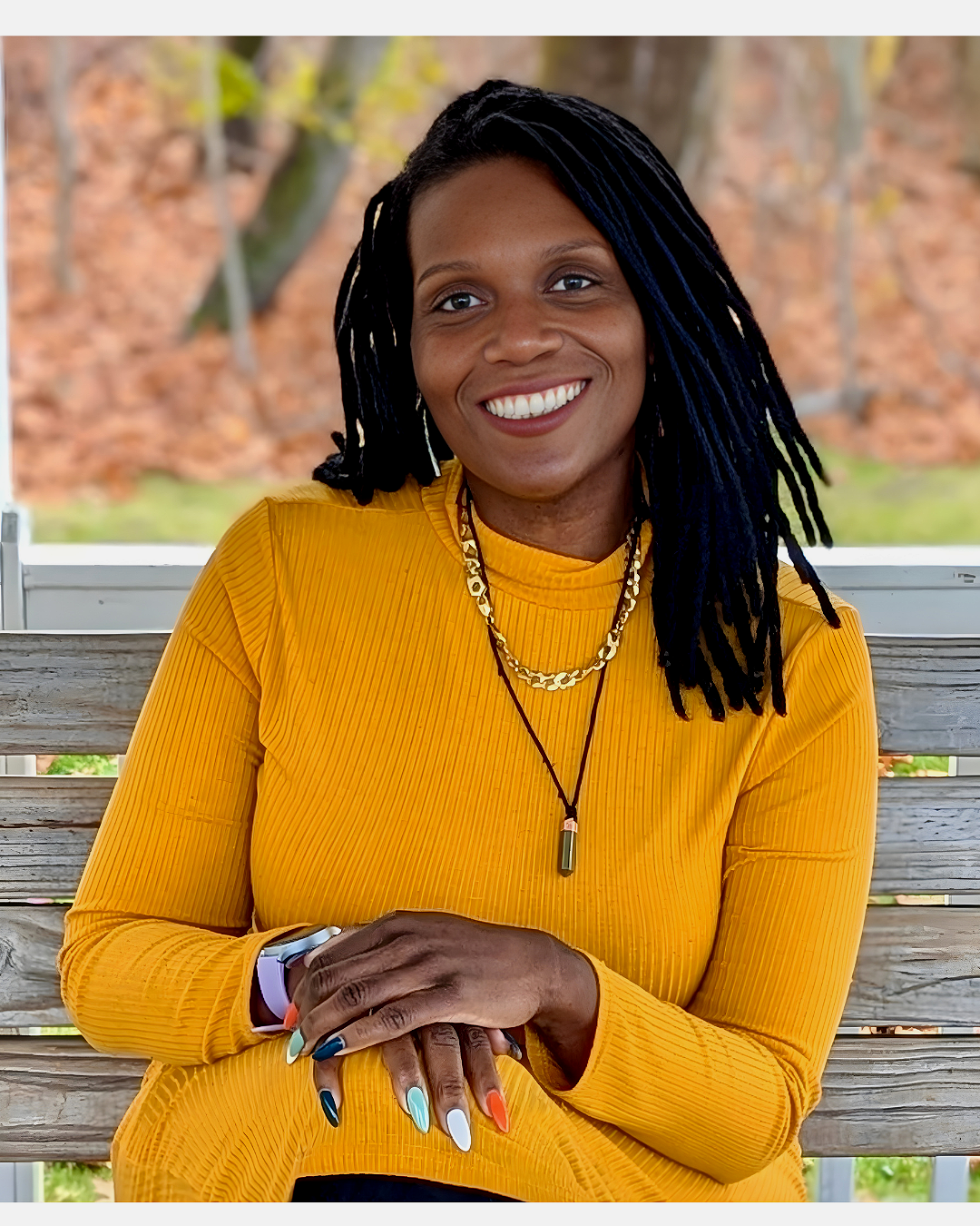 A woman with dark styled dreadlocks, smiling in a yellow ribbed top, sitting outdoors on a wooden bench with trees and fall foliage in the background.